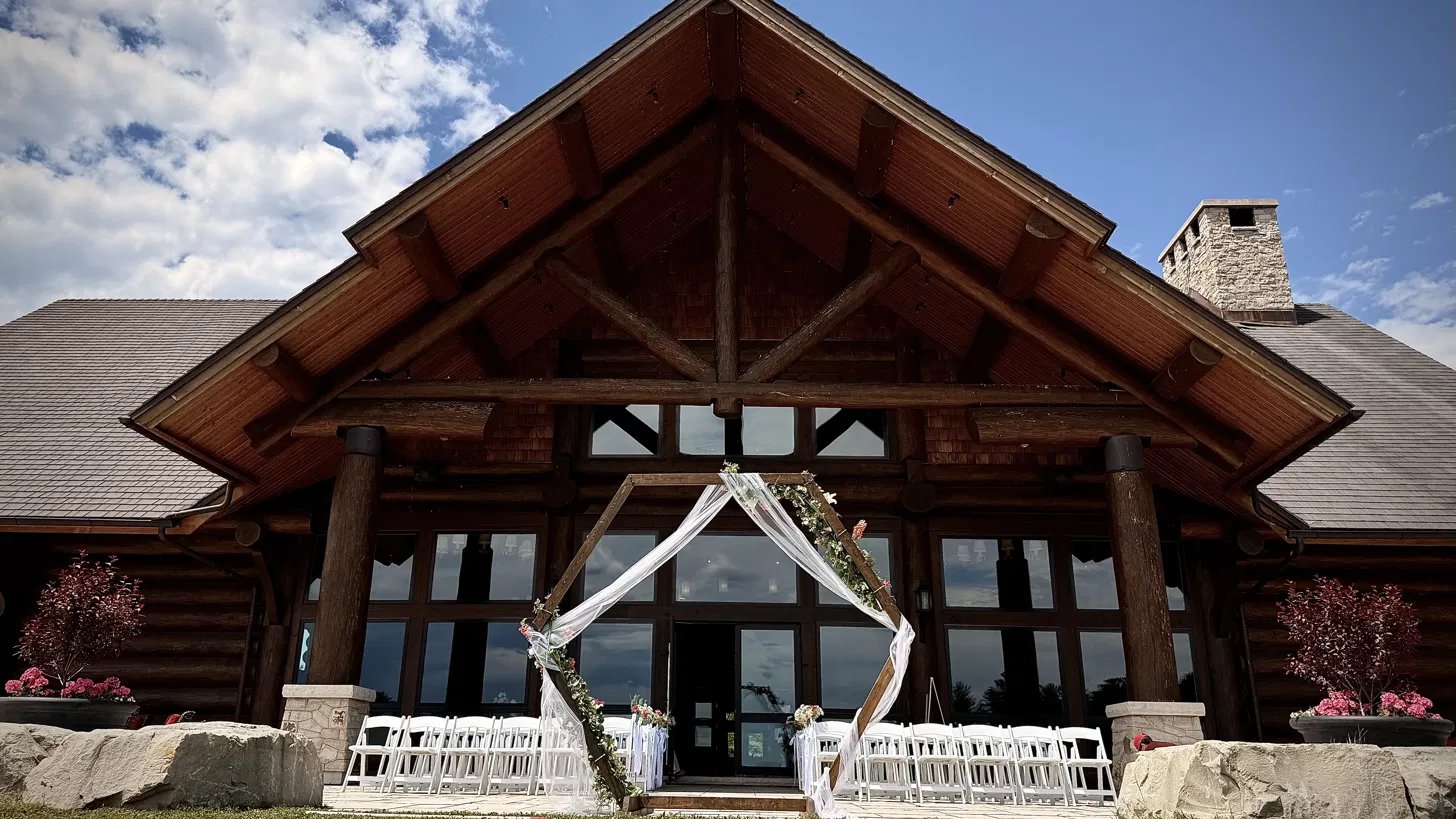 A hexagonal floral arch and white ceremony chairs before the great log gable of Canada House