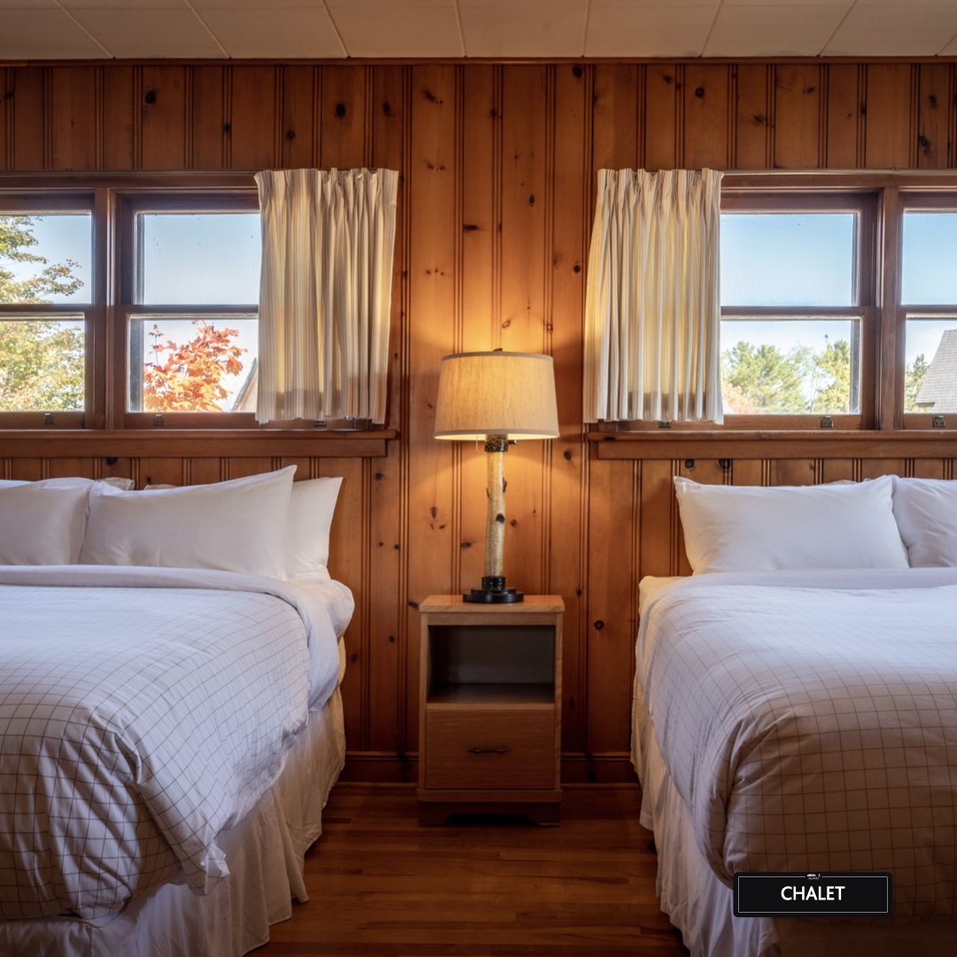 Pine-panelled chalet bedroom with two beds and fall foliage through the windows