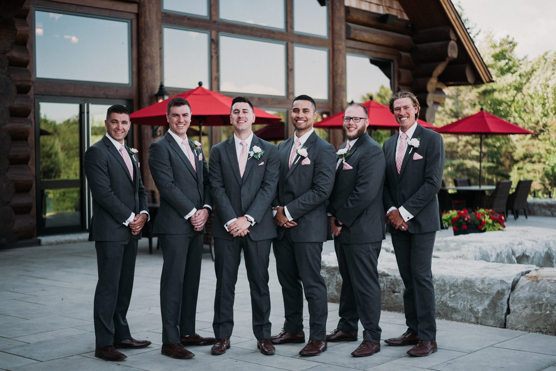 Six groomsmen in grey suits and pink ties in front of the log lodge